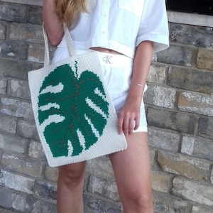 May include: A woman is holding a white crocheted tote bag with a green monstera leaf design. The bag has a long strap and is worn over the shoulder. The woman is wearing a white shirt and white shorts.