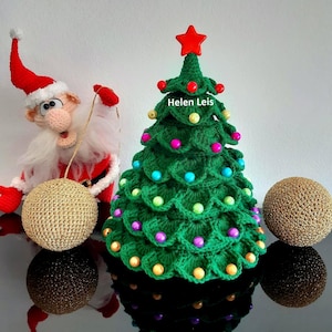 May include: A green crocheted Christmas tree with colorful beads and a red star on top. A crocheted Santa Claus figure is holding a gold ball ornament. The tree has a sign that says "Helen Leis".