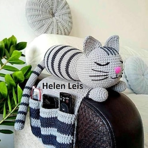 以下が含まれることがあります： グレーと黒のストライプの猫型かぎ針編みオーガナイザー。濃い茶色の表面に置かれています。猫はピンクの鼻と閉じた目をしています。オーガナイザーには小物を収納できるポケットが付いています。「Helen Leis」の文字が見えます。