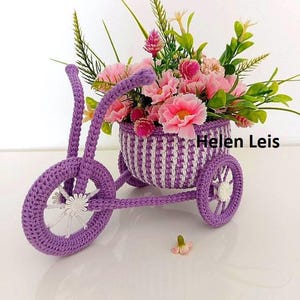 Puede incluir: Un jardinera de triciclo morado tejido a ganchillo, llena de flores rosas y vegetación. La cesta está rayada con blanco y morado. Las ruedas tienen detalles blancos. El texto "Helen Leis" es visible.