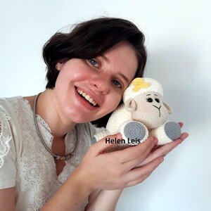 May include: A woman is holding a crocheted white sheep with a yellow flower on its head. The sheep has grey feet and a crocheted nose. The woman is smiling and the text "Helen Leis" is visible in the image.
