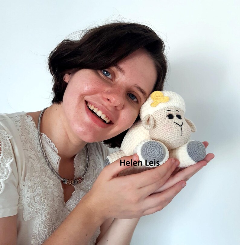 May include: A woman is holding a crocheted white sheep with a yellow flower on its head. The sheep has grey feet and a crocheted nose. The woman is smiling and the text "Helen Leis" is visible in the image.