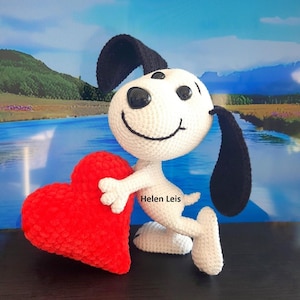Può includere: Un cane bianco all'uncinetto che tiene un giocattolo di peluche a forma di cuore rosso. Il cane ha le orecchie nere e un naso nero. Il testo "Helen Leis" è visibile sul corpo del cane.