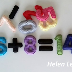 May include: Crocheted felt numbers in various colors, including pink, blue, green, purple, and black, spelling out the equation "6 + 8 = 14". The numbers are arranged in a line, with the plus sign, equals sign, and the number 14 in between. The numbers are all crocheted and have a 3D appearance. The image also includes the text "Helen Leis" at the bottom.