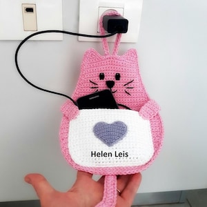 以下が含まれることがあります： ピンク色の猫の形をしたかぎ針編みの携帯電話ホルダー。白いポケットと紫色のハートのデザイン。ホルダーは、コンセントに差し込まれた黒い充電器に接続されています。携帯電話はポケットの中にあります。