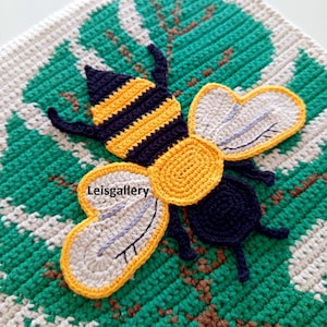 Puede incluir: Cuadrado de ganchillo hecho a mano con un diseño de abeja. La abeja tiene rayas amarillas y negras, con alas blancas y grises. El fondo es verde y blanco, con el texto "Leisgallery" visible.