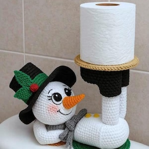 May include: A handmade crochet snowman toilet paper holder. The snowman has a black top hat with green holly leaves and a red berry, an orange carrot nose, and a gray scarf. The holder is white, black, and green.