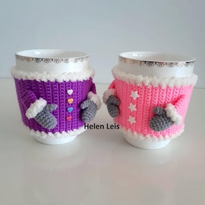 Puede incluir: Dos calientavasos de ganchillo, uno morado con botones de corazón de colores y otro rosa con botones de estrella blanca. Ambos calientavasos tienen guantes de ganchillo grises y ribete de piel sintética blanca. El texto "Helen Leis" es visible en la imagen.