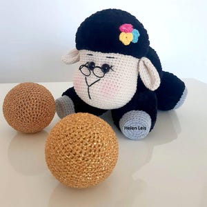 May include: A black and white crocheted sheep toy with a pink nose and black eyes. The sheep is wearing glasses and has a colourful flower on its head. The sheep is sitting on a white surface with two gold balls.