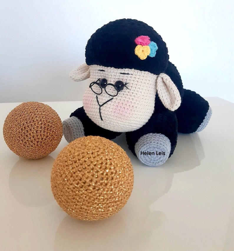 May include: A black and white crocheted sheep toy with a pink nose and black eyes. The sheep is wearing glasses and has a colourful flower on its head. The sheep is sitting on a white surface with two gold balls.