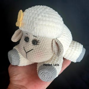 May include: A crocheted white sheep with grey hooves and a yellow flower on its head. The sheep is lying on its side with its head turned to the side. The sheep is a handmade toy.