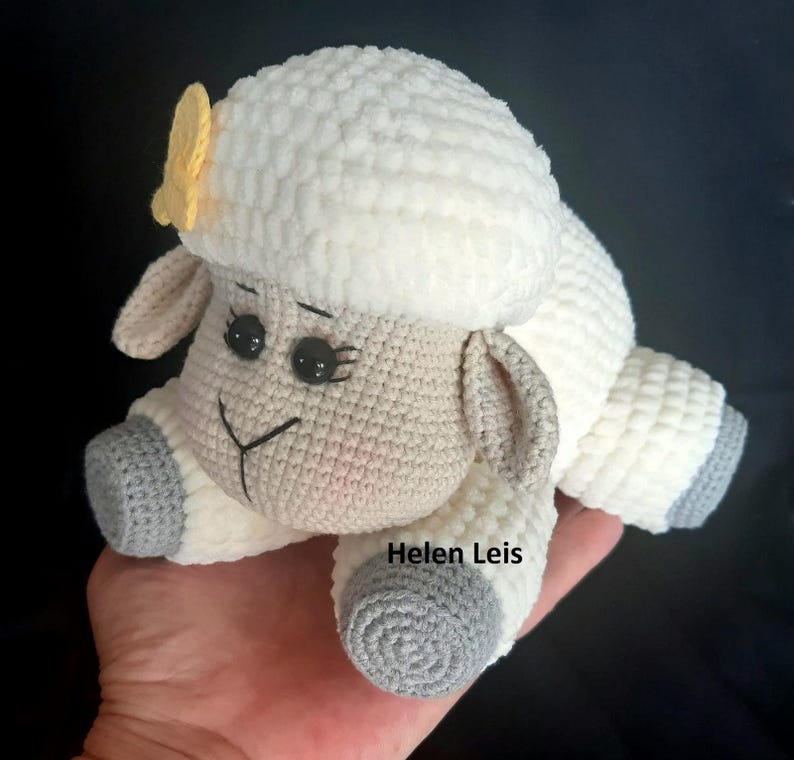 May include: A crocheted white sheep with grey hooves and a yellow flower on its head. The sheep is lying on its side with its head turned to the side. The sheep is a handmade toy.