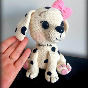 Puede incluir: Un perro dálmata de ganchillo hecho a mano con manchas negras, ojos grandes y negros y un lazo rosa. El perro tiene detalles en rosa en las patas y las mejillas. El perro está sentado sobre una superficie negra.