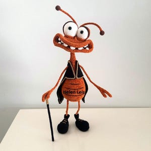 May include: A crocheted orange and brown bug with large white eyes and a toothy grin. The bug is wearing a black vest with gold trim and is holding a black walking stick. The bug is standing on a white surface. The text "Helen Leis" is embroidered on the vest.