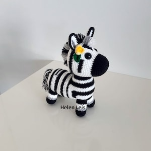 May include: A handmade crochet zebra figurine with black and white stripes. The zebra has a black mane, tail, and nose, with a yellow flower and green leaves on its head. The figurine is standing on four small legs.