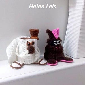 Op de afbeelding: Twee handgemaakte gehaakte figuren. Eén is wit met een bruine hoge hoed en sandalen, de andere is bruin met een roze strik en voeten. De figuren hebben grote ogen en zitten op een wit oppervlak. De tekst op de muur luidt "Helen Leis".