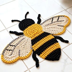 Puede incluir: Una alfombra de ganchillo con forma de abeja en amarillo, negro y crema. La abeja tiene un cuerpo amarillo con rayas negras, alas color crema y antenas y patas negras. La alfombra está colocada sobre una superficie de baldosas blancas.