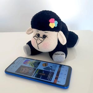 May include: A black and white crocheted sheep with a pink, blue and yellow flower on its head. The sheep is wearing glasses and is sitting on a table next to a blue smartphone.