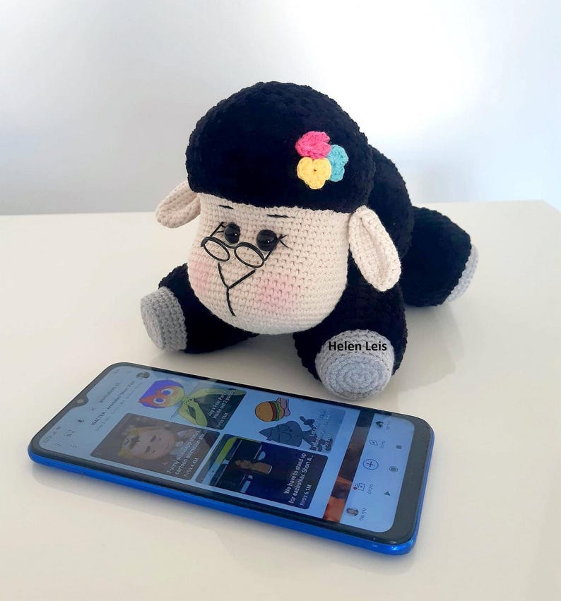 May include: A black and white crocheted sheep with a pink, blue and yellow flower on its head. The sheep is wearing glasses and is sitting on a table next to a blue smartphone.