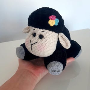 May include: A black and white crocheted sheep plush toy with a flower on its head. The sheep is sitting on a hand.
