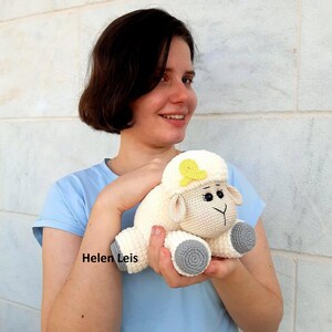 May include: A white crocheted sheep plush toy with a yellow bow on its head and grey feet. The sheep has black eyes and a black nose.