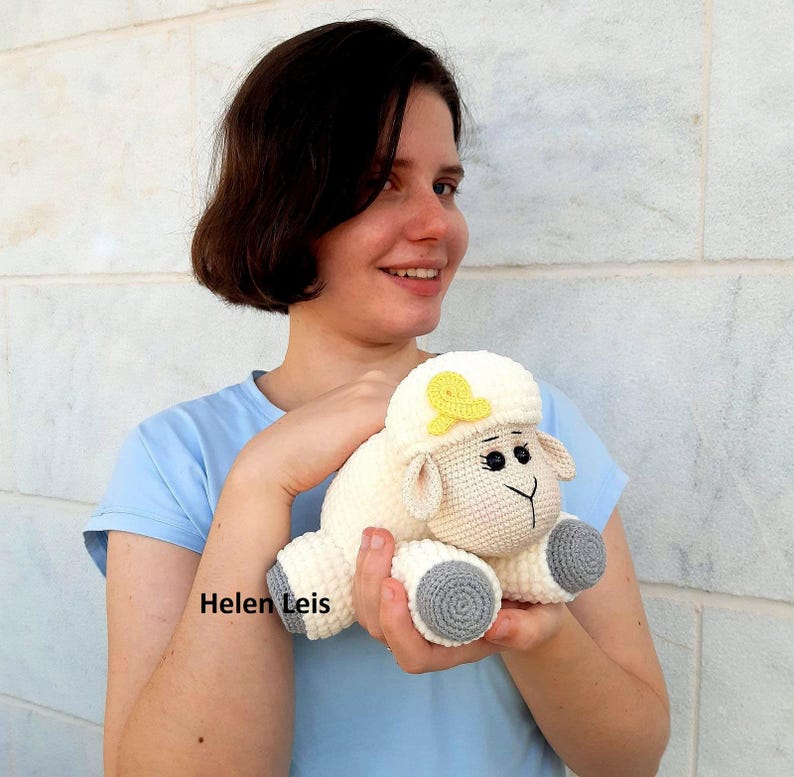 May include: A white crocheted sheep plush toy with a yellow bow on its head and grey feet. The sheep has black eyes and a black nose.