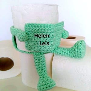 May include: A crocheted green toilet paper holder shaped like a character, with the name "Helen Leis" on the front. The holder is wrapped around a roll of white toilet paper, with additional rolls in the background.