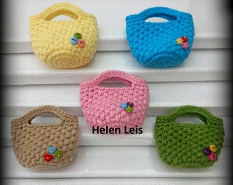 Op de afbeelding: Vijf miniatuur gehaakte handtassen in geel, blauw, roze, bruin en groen. Elke tas heeft een handvat en is versierd met kleurrijke knopen. De tekst "Helen Leis" is zichtbaar op de roze tas.