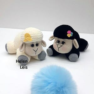 May include: Two crocheted sheep plushies, one white with a yellow bow and one black with colourful flowers. The sheep are lying on a white surface with a blue fluffy ball in the foreground.