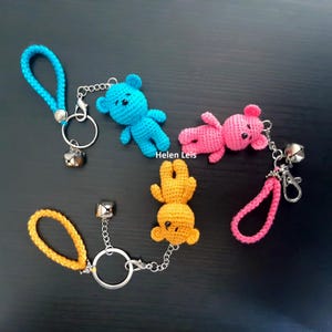 Puede incluir: Tres llaveros de osito de peluche tejidos a crochet con cascabeles. Uno es azul, otro rosa y otro naranja.