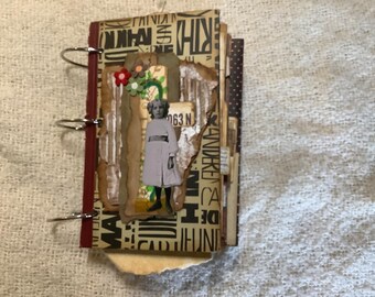 Op de afbeelding: Een klein ringband-plakboek met een beige kaft met zwarte typografie, een centraal collage-element met een vintage-stijl foto van een kind in een witte jurk, gelaagd met golfkarton en kleine rode, groene en bruine bloemaccenten.