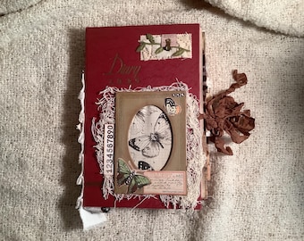 Op de afbeelding: Een handgemaakt rood dagboek met een vlinderontwerp. Het dagboek heeft een omlijste ovale afbeelding van een vlinder, omgeven door decoratieve elementen en het woord "Diary" op de omslag. Het dagboek is versierd met kant en andere decoratieve elementen.