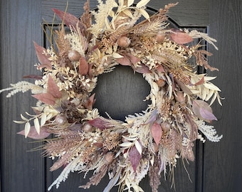 May include: A dried floral wreath with a variety of textures and colors. The wreath features beige, cream, and mauve-colored dried flowers, leaves, and seed pods. It is hanging on a black door.