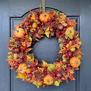 May include: A fall-themed wreath with pumpkins, berries, pinecones, and colorful leaves. The wreath is arranged in a circular shape and is attached to a dark blue door. The wreath features orange, red, yellow, and green colors.
