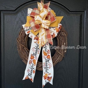 May include: A grapevine wreath adorned with a large bow featuring autumn colors. The bow has layers of orange, yellow, and plaid ribbons. Two long white ribbons with the word "Thankful" and leaf designs hang down. The text "Cotton Blossom Decor" is visible.