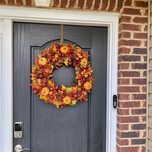 May include: A fall-themed wreath with pumpkins, berries, and leaves hangs on a black door. The wreath features orange, red, and yellow colors, creating a festive autumn display. The door is framed by a brick wall.