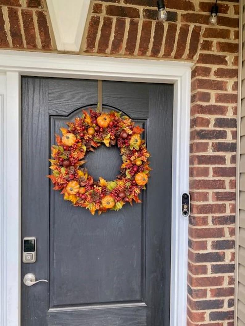 May include: A fall-themed wreath with pumpkins, berries, and leaves hangs on a black door. The wreath features orange, red, and yellow colors, creating a festive autumn display. The door is framed by a brick wall.