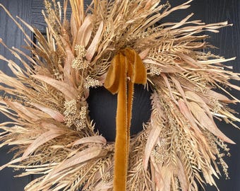 May include: A dried floral wreath with a variety of beige and tan grasses, leaves, and small flowers. A long, mustard-colored velvet ribbon hangs from the wreath. The wreath is displayed on a dark blue door.