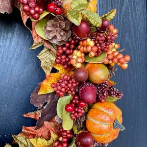 May include: A fall-themed wreath with pumpkins, berries, and leaves in shades of orange, red, and brown. The wreath includes pinecones and faux fruit, creating a festive autumn decoration.