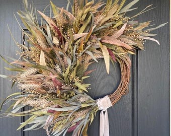 May include: A rustic wreath made of dried wheat, leaves, and other natural elements. The wreath features a variety of colors, including green, brown, and pink, and is tied with a cream-colored ribbon. This decorative wreath is perfect for adding a touch of nature to any home.
