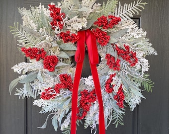 May include: A festive Christmas wreath with red berries and a red velvet ribbon. The wreath is made of faux greenery with a frosted appearance. The wreath is displayed on a black door.