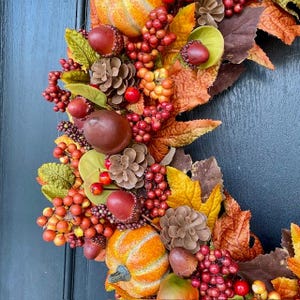 May include: A fall-themed wreath with pumpkins, pinecones, berries, and leaves in shades of orange, red, brown, and green. The wreath is a decorative item for the autumn season, suitable for a door or wall.
