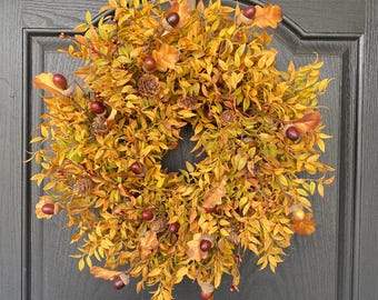May include: A circular autumn wreath with yellow and orange leaves, acorns, and small pinecones. The wreath is hanging on a dark gray door, creating a warm, seasonal decoration. The wreath is a festive decoration for the fall season.
