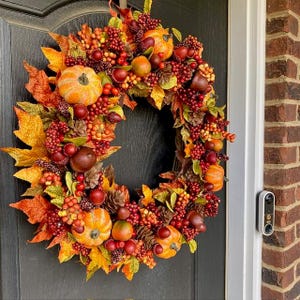 May include: A fall-themed wreath with pumpkins, berries, and leaves in shades of orange, red, and yellow. The wreath is adorned with small pumpkins, pinecones, and various fruits, creating a festive and seasonal decoration.
