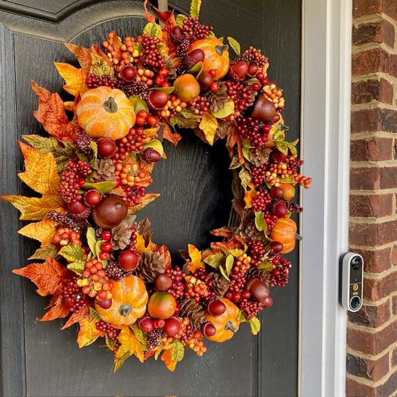May include: A fall-themed wreath with pumpkins, berries, and leaves in shades of orange, red, and yellow. The wreath is adorned with small pumpkins, pinecones, and various fruits, creating a festive and seasonal decoration.