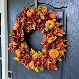 May include: A fall-themed wreath with pumpkins, berries, and leaves in shades of orange, red, and green. The wreath is displayed on a dark blue door, creating a warm and inviting autumn decoration.