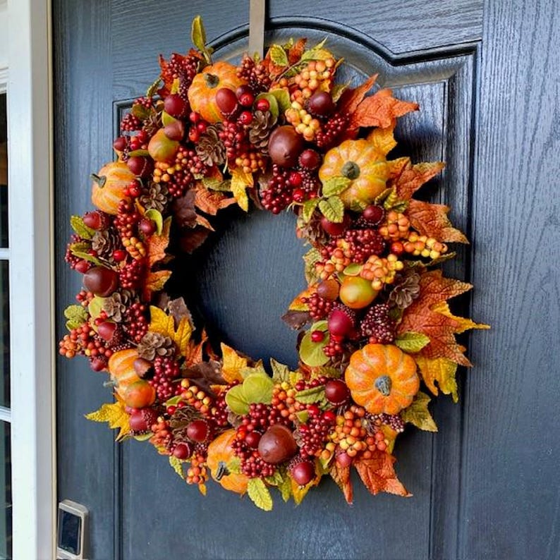 May include: A fall-themed wreath with pumpkins, berries, and leaves in shades of orange, red, and green. The wreath is displayed on a dark blue door, creating a warm and inviting autumn decoration.