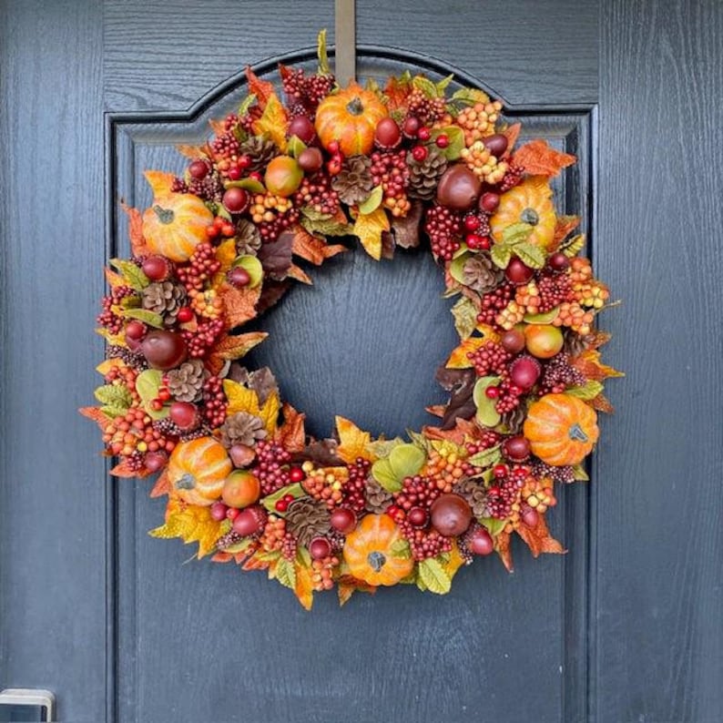 May include: A fall-themed wreath with pumpkins, berries, pinecones, and colorful leaves. The wreath is arranged in a circular shape and is attached to a dark blue door. The wreath features orange, red, yellow, and green colors.