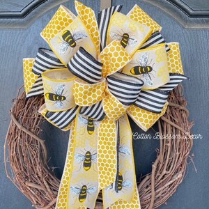 May include: A wreath with a large yellow and black bow with a bee pattern. The bow is tied with a black and white striped ribbon. The wreath is made of grapevine and has a brown finish. The text "Cotton Blossom Decor" is visible in the bottom right corner of the image.
