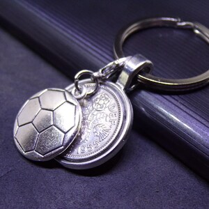 Könnte beinhalten: Silberner Schlüsselanhänger mit einem Fußball-Anhänger und einer Vintage-Münze. Der Fußball-Anhänger hat ein detailliertes sechseckiges Muster. Die Münze zeigt ein Blumendesign und das Jahr 1955.